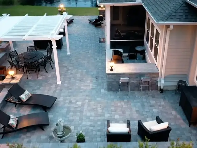 A large gray paver patio at dusk seen from above, wrapping around a house, with a white pergola over a dining set, loungers and lit lanterns on the seating wall columns