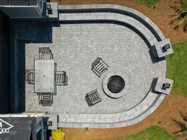 An overhead view of a horseshoe-shaped paver patio, a light gray field inside a charcoal border, wrapped by a curved seating wall with four capped columns and a round fire pit at the head