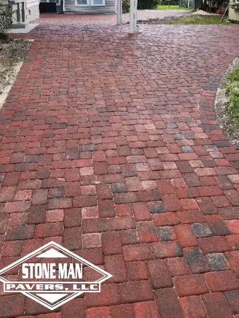 A broad driveway of tumbled clay pavers in mottled reds and browns, laid in running bond and running away between a house and a shrub bed toward a white-posted porch