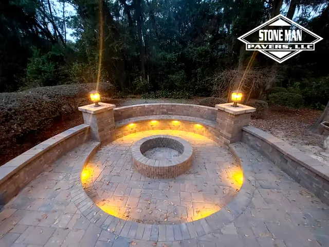 A round paver fire pit at the center of a circular paver patio at dusk, with a curved seating wall lit from under its cap and two capped columns carrying lit lanterns, under live oaks hung with Spanish moss