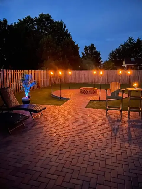 A red-toned paver patio after dark with a round fire pit at its center, Adirondack chairs and a lounger around it, and string lights run along a wood fence