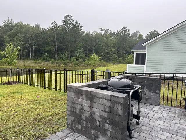 A gray block grill counter with a ceramic kamado cooker and a gas grill set in, standing at the edge of a gray paver patio behind a house, with a black aluminum fence and a treeline beyond