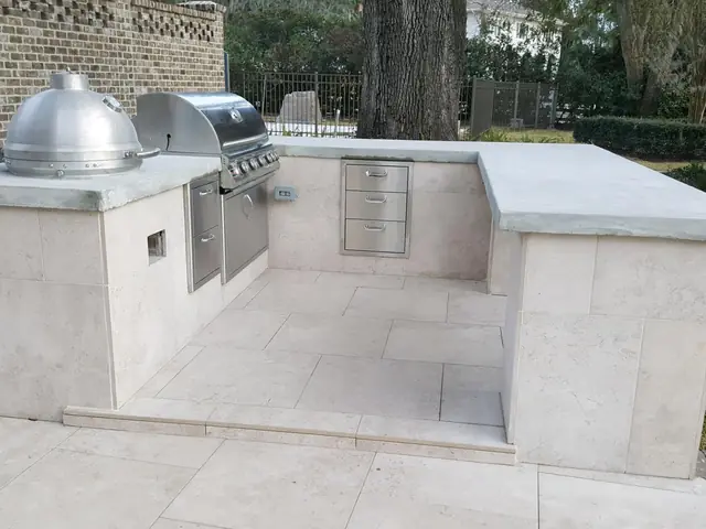 A U-shaped outdoor kitchen clad in cream stone tile with a gray poured countertop, a stainless gas grill and a ceramic kamado cooker set into it