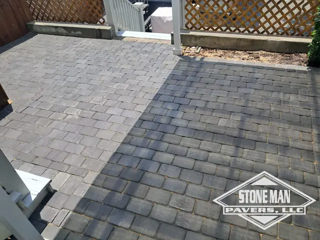 A back patio of gray tumbled pavers running up to a white-railed deck and a lattice screen, with a charcoal soldier course along the near edge and a mulch bed behind