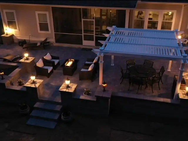 A raised paver patio at night behind a house, with a white pergola over a dining set, wicker seating around a linear fire table, and capped columns along the seating wall each carrying a lit lantern