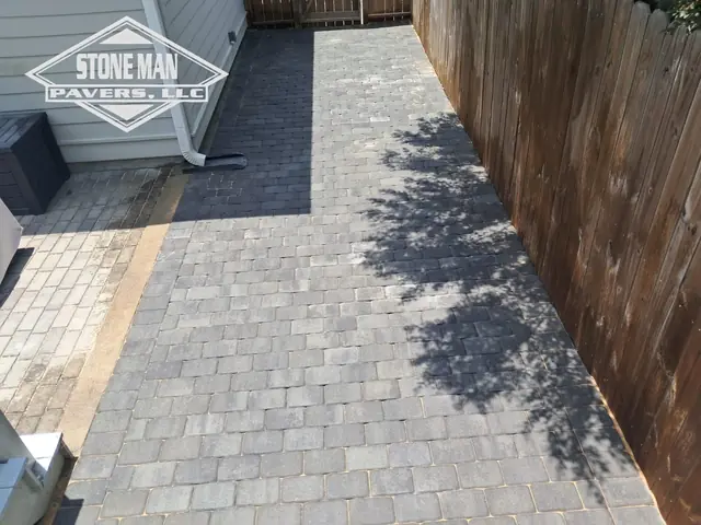 A run of gray tumbled pavers between a clapboard house wall and a tall wood privacy fence, with a charcoal band across the far end and cream stone paving at the near corner