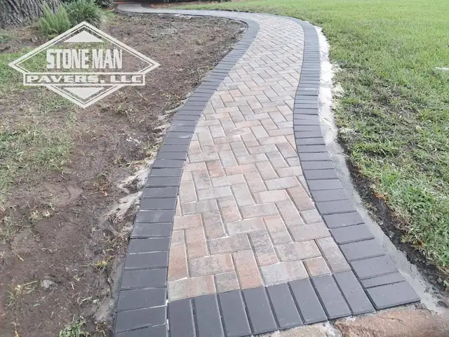 The finished walk: clay-toned pavers laid in herringbone inside a charcoal soldier-course border, curving from the curb up toward the house, with bare soil either side where the trench was