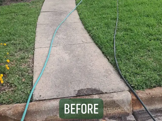 A narrow poured concrete walk, stained and worn, curving from the street curb up to the door of a brick ranch house, with garden hoses lying across it