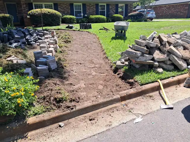 The old concrete walk broken out and stacked in slabs on the lawn beside pallets of new pavers, with a wheelbarrow and an open trench where the walk was