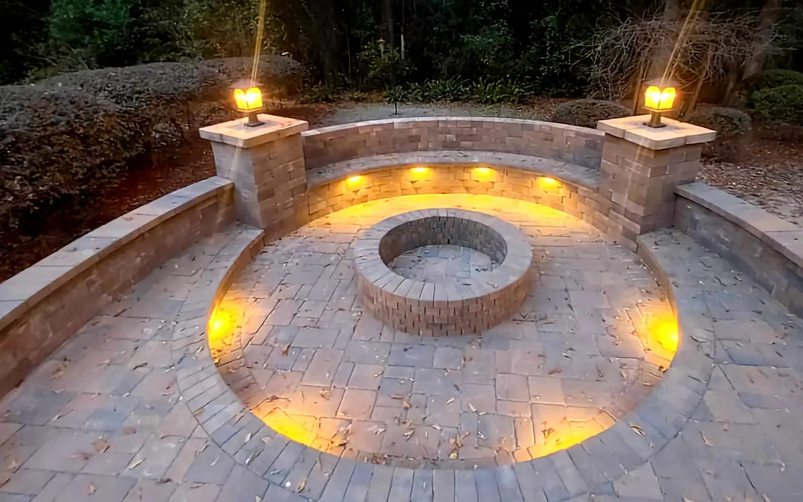 A round paver fire pit at the center of a circular paver patio at dusk, with a curved seating wall lit from under its cap and two capped columns carrying lit lanterns, under live oaks hung with Spanish moss